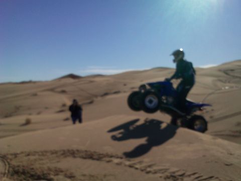 2004 Jeep Wrangler - A person in a helmet is riding a blue ATV, jumping over a sand dune. Another person stands in the background on a sandy landscape under a c…