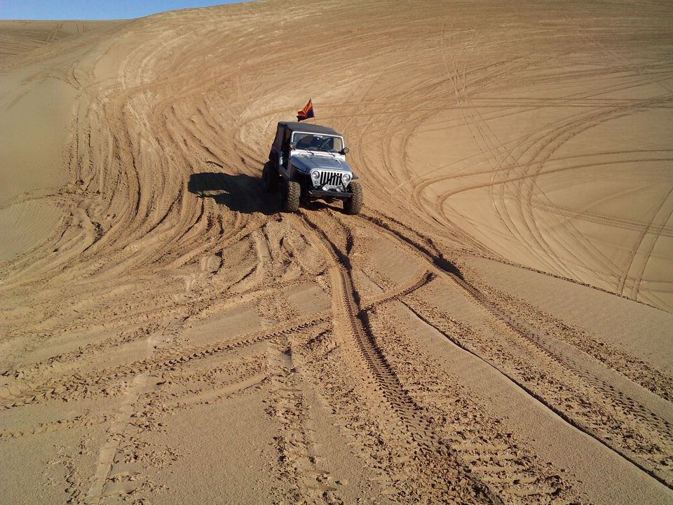 Silver Jeep Wrangler driving on a sand dune with tire tracks.