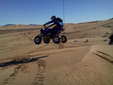 2004 Jeep Wrangler - A person on a blue ATV is airborne over sand dunes, performing a jump. The ATV is mid-air with its wheels off the ground, and the rider is…