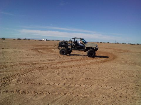 2004 Jeep Wrangler - A custom-built off-road buggy with a roll cage is shown performing a sharp turn on a sandy, desert-like terrain. The vehicle has large, agg…