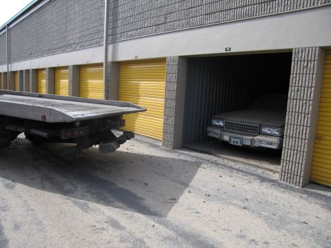 1991 Ford LTD Crown Victoria - A tow truck is positioned next to a storage unit where a dusty 1987 Chevrolet Caprice Classic is parked.