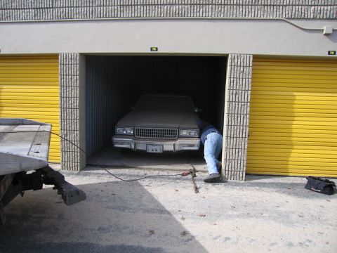 1991 Ford LTD Crown Victoria - A person is pushing a dusty 1986 Chevrolet Caprice out of a storage unit with yellow doors, while a tow truck is attached.