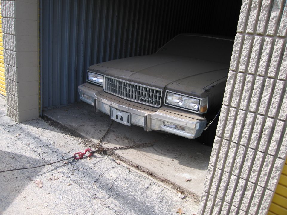 Dusty car, possibly 1980s Chevrolet Caprice, parked in a garage with a chain attached to the front bumper