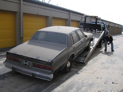 1991 Ford LTD Crown Victoria - A dusty Chevrolet Caprice sedan is loaded onto a tow truck in front of storage units.