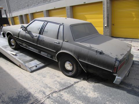 1991 Ford LTD Crown Victoria - A heavily soiled sedan is being loaded onto a tow truck, its dark paint obscured by a thick layer of dust.