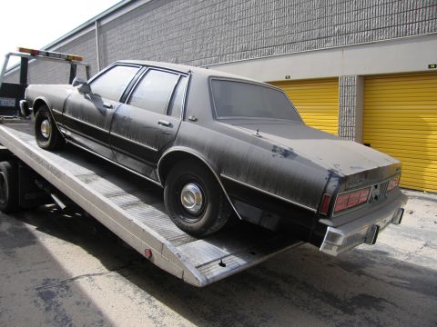 1991 Ford LTD Crown Victoria - A dusty dark sedan is loaded onto a tow truck, showing its rear profile with chrome accents and rectangular taillights.