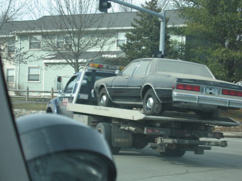 1991 Ford LTD Crown Victoria - A tow truck transports a dark-colored sedan on its flatbed, seen from the side.