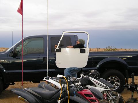 1986 Toyota Hilux - A person sits in the driver's seat of a Ford F-350 truck, with a Jeep door propped open, parked next to ATVs in a sandy area.