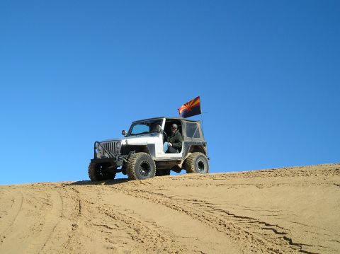 1986 Toyota Hilux - A Jeep Wrangler with an Arizona flag flies over a sand dune under a clear blue sky.