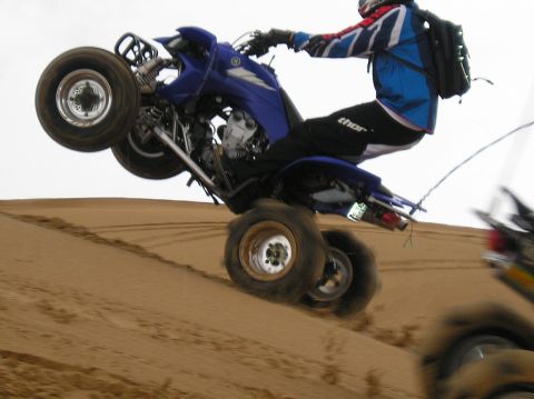 1986 Toyota Hilux - A rider performs a wheelie on a blue quad bike while navigating sand dunes, with another vehicle visible in the background.
