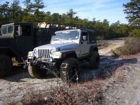 2004 Jeep Wrangler - A silver Jeep Wrangler is parked on a dirt road next to a military truck carrying lumber.