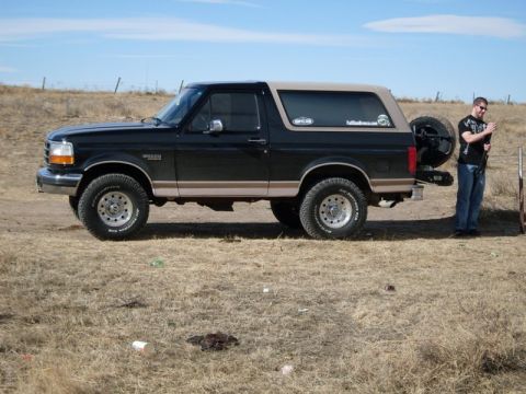 1996 Ford Bronco - A black 1996 Ford Bronco with a tan removable top is parked outdoors on a dry, grassy field. A man stands near the rear of the vehicle.