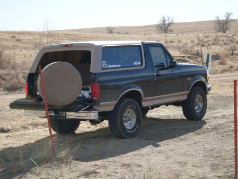 1996 Ford Bronco - Black and tan two-tone Ford Bronco shot from the rear three-quarter on a dirt track in open dry grassland, tailgate down with the glass up…