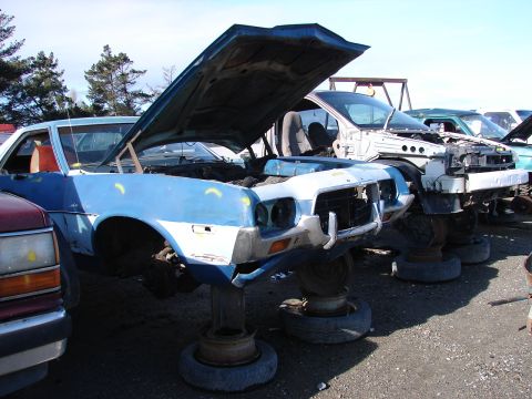 1973 Ford Ranchero - A stripped blue and white 1970s Ford body sits propped on stacked wheels in a salvage yard, hood up, front fenders and quarter panel wearin…