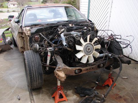 1973 Ford Ranchero - A faded gold 1980s Ford coupe sits on jack stands with the hood, fenders, radiator and front bodywork stripped off, exposing the V8, white…