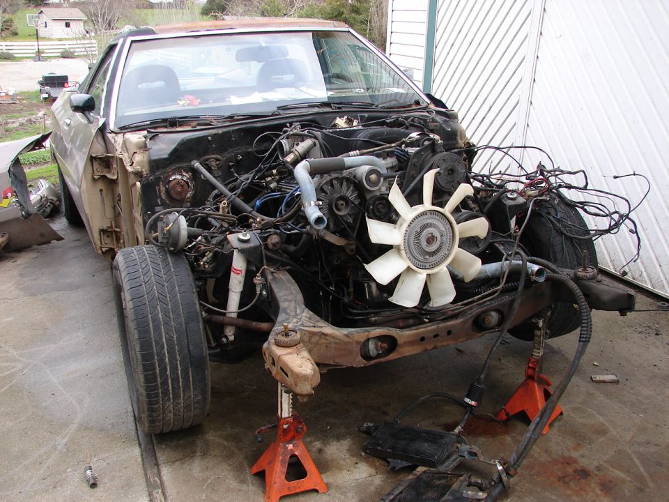 Gold 1980s Ford coupe on jack stands with front end stripped, exposing V8 and wiring