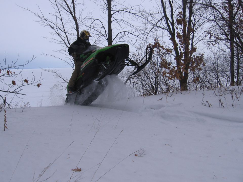 Green and black Arctic Cat snowmobile airborne over snow with rider, bare trees behind