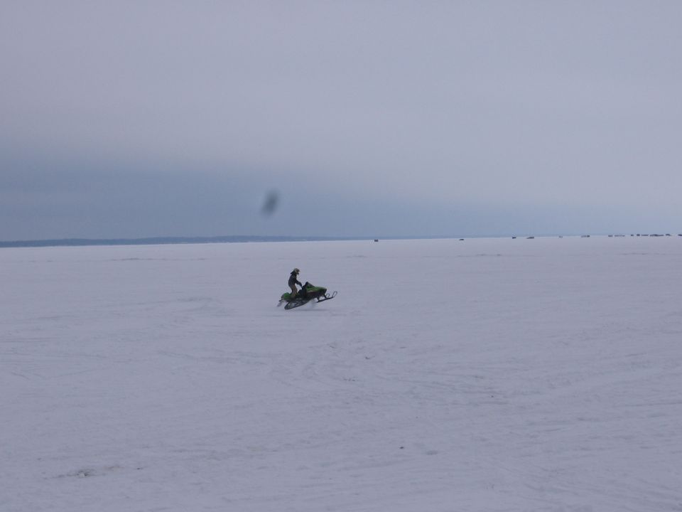 Green snowmobile riding wheelie across a frozen lake under overcast sky