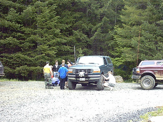 Lifted teal Ford Bronco with winch bumper on gravel trailhead, people gathered around