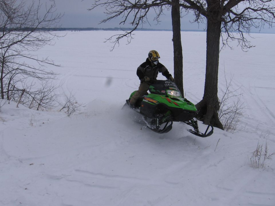 Rider on a green snowmobile catching air near a tree on a snowy lakeshore