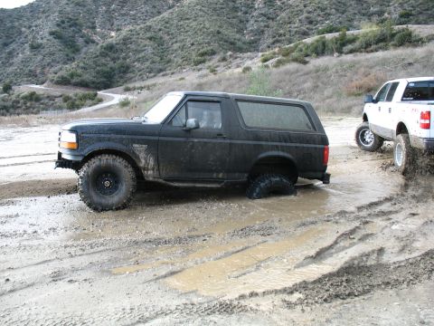 1987 Ford Bronco - A black Ford Bronco navigates a muddy off-road trail, with a white pickup truck following closely behind.