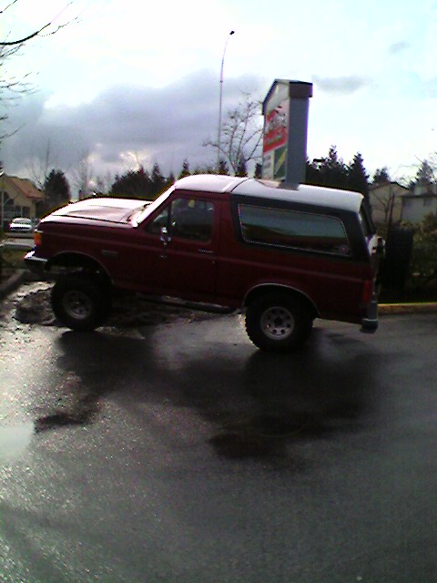 1989 Ford Bronco - A red Ford Bronco with a black top is parked on a wet surface under a cloudy sky.
