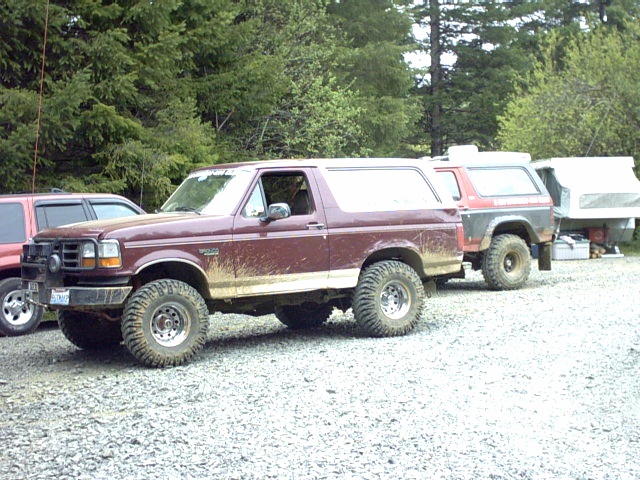Lifted maroon Ford Bronco with mud-splattered sides on a gravel lot, another Bronco and camper behind
