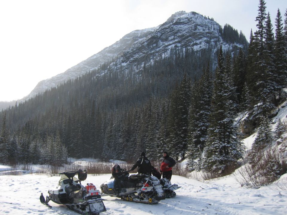 Two snowmobiles parked in snow with people standing near them; a large, snow-covered mountain and pine forest in the background