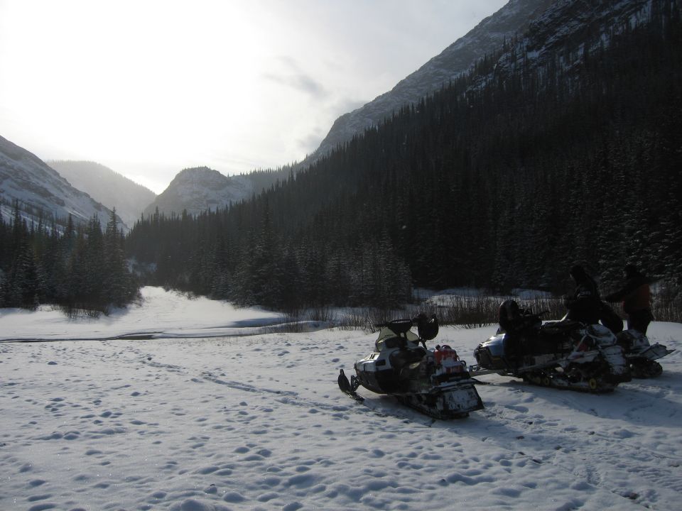 Two snowmobiles parked in a snowy valley with mountains and trees in the background