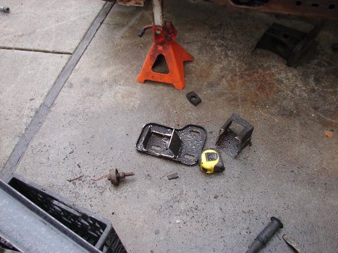 1973 Ford Ranchero - Removed suspension and mount hardware laid out on a stained concrete garage floor: a black crossmember mount bracket, a small welded box br…