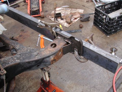 1973 Ford Ranchero - Bare vehicle frame under repair on a concrete garage floor, with a new length of rectangular tube steel clamped in place by an orange bar c…