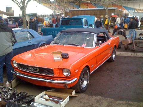 1983 Ford Bronco - Orange 1965 Ford Mustang convertible at an outdoor car show with other vehicles and parts visible.