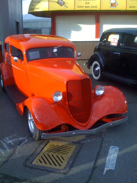 1983 Ford Bronco - An orange 1933 hot rod coupe is parked next to a black car in front of a building with a yellow awning.
