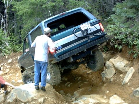 1995 Ford Bronco - A teal 1995 Ford Bronco is shown navigating a challenging rocky off-road trail with assistance from a person pushing.