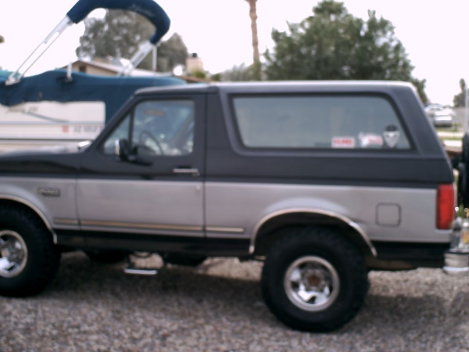 Side view of a lifted two-tone black and silver Ford Bronco SUV on gravel with a boat in the background