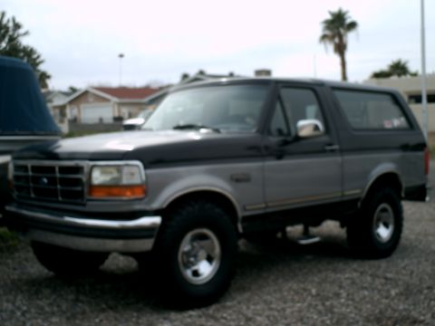 1994 Ford Bronco - A two-tone black and silver Ford Bronco is parked on a gravel lot with houses and a palm tree in the background. The SUV is viewed from a 3…
