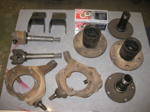1979 Ford Bronco - A collection of rusty 1979 Ford Bronco front axle and hub components are laid out on a gray surface. The parts include steering knuckles, a…