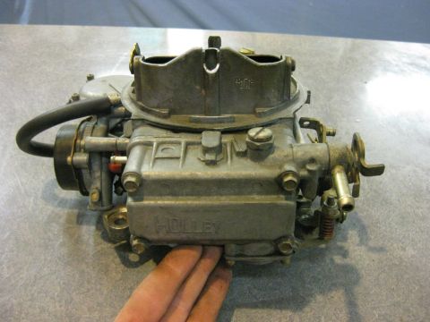 1979 Ford Bronco - A Holley carburetor is shown from a 3/4 front angle, resting on a speckled gray surface. A hand is visible at the bottom of the frame, hold…
