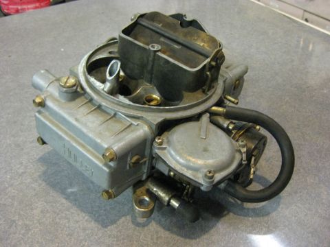 1979 Ford Bronco - A Holley carburetor is shown from a 3/4 front angle, resting on a speckled gray surface. The carburetor is silver and black with a black ho…
