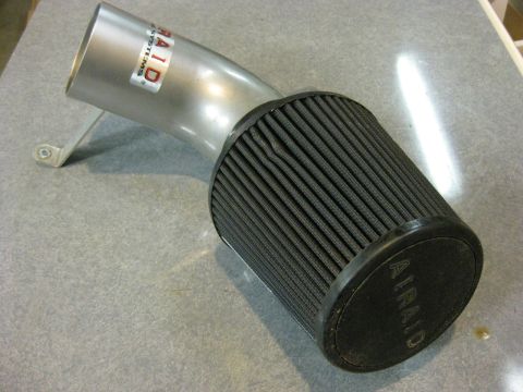 1979 Ford Bronco - A silver Airaid cold air intake system with a black filter is shown on a speckled gray surface. The intake tube has a red and white Airaid…
