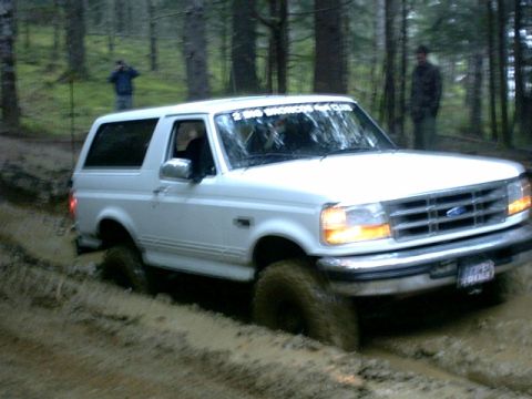 1995 Ford Bronco - A white Ford Bronco navigates a muddy trail in a forest, with two people visible in the background.