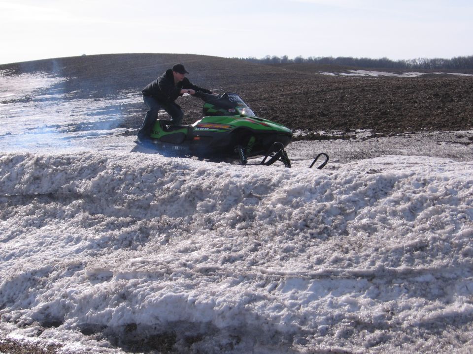 Man riding a green Arctic Cat snowmobile through slushy snow beside a bare field