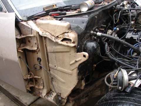 1973 Ford Ranchero - Engine bay of a car with the front fender removed, exposing a rusty tan inner fender apron with a rotted-through hole, the hinge pillar and…