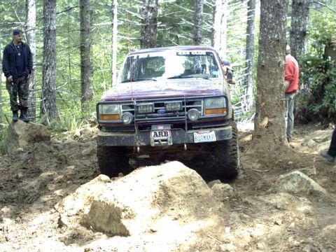 1995 Ford Bronco - Ford Bronco tackles a challenging off-road course in a wooded area, with spectators nearby.