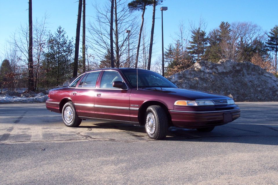 Maroon 2001 Ford Crown Victoria sedan parked on asphalt with snow pile and trees in background.