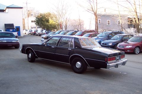 2001 Ford Crown Victoria - A black 1980s sedan is parked in a lot with other vehicles. The sedan has chrome trim and a chrome bumper.