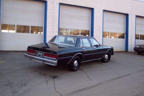 2001 Ford Crown Victoria - A black 1980s Chevrolet Caprice sedan is parked in front of a building with three garage doors. The car is viewed from the rear-left quarte…