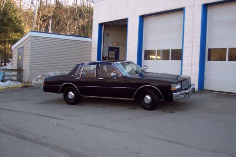 2001 Ford Crown Victoria - A black four-door sedan is parked in front of a building with two large garage doors. The car is viewed from the side at a slight front ang…