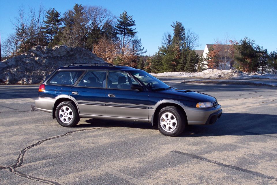 Blue and silver Subaru Outback wagon parked on asphalt with snow pile in background.