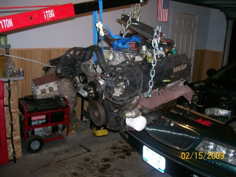 1995 Mercury Grand Marquis - A V8 engine is suspended by a red engine hoist and chains over the front end of a dark green sedan. A red Craftsman generator is visible to…